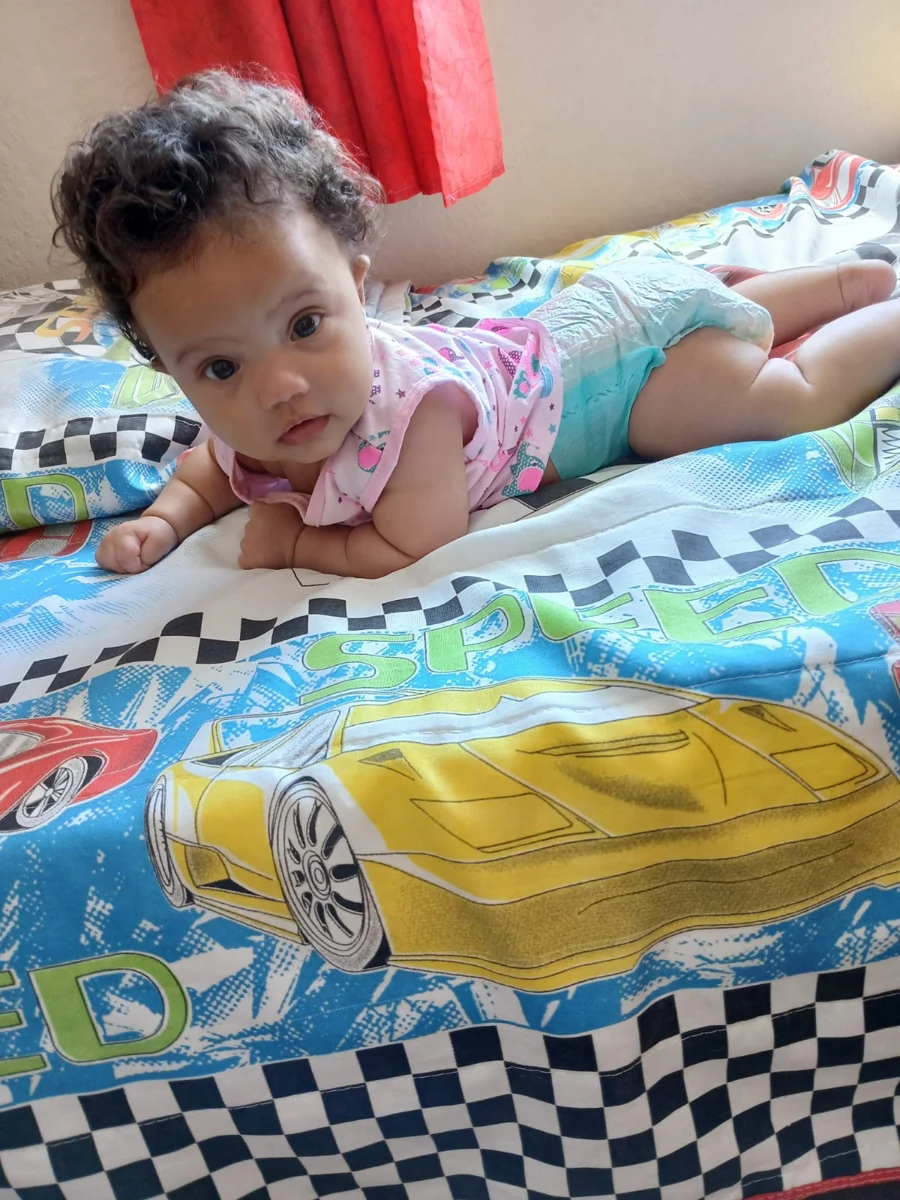 Bebê apoiada na cama, na foto de referência enviada.